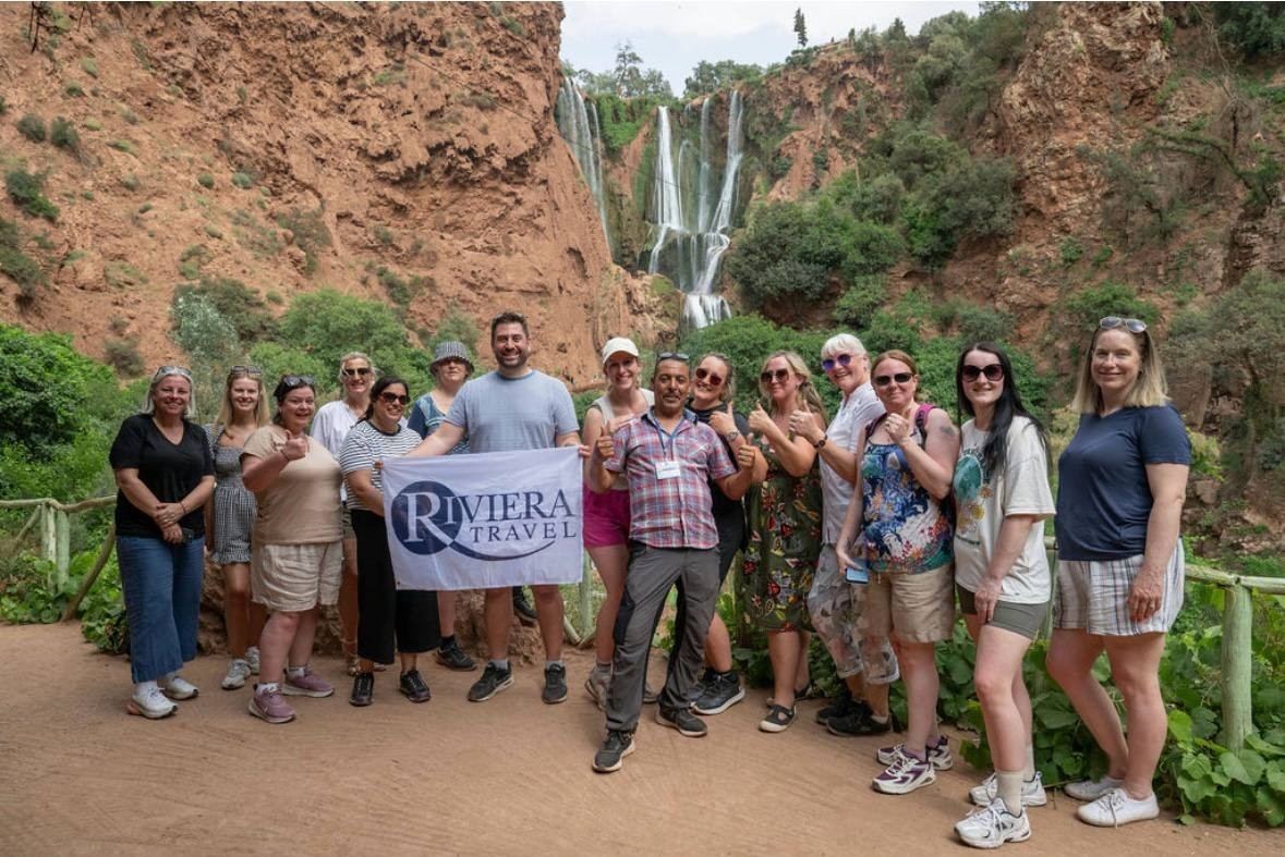 A group in front of the waterfalls, holding a Riviera Travel flag